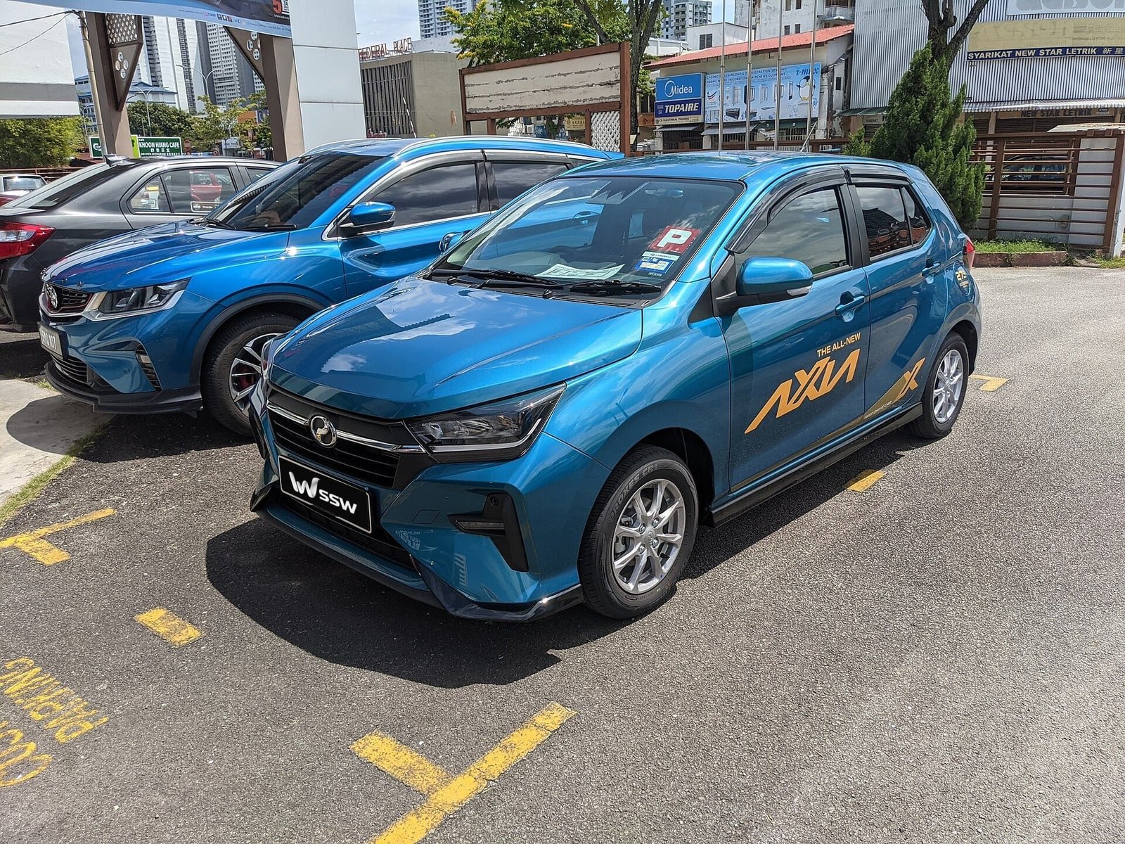 Coral blue Perodua Axia 1.0 AV hatchback, front three-quarter view in a sunlit forecourt in Penang, Malaysia