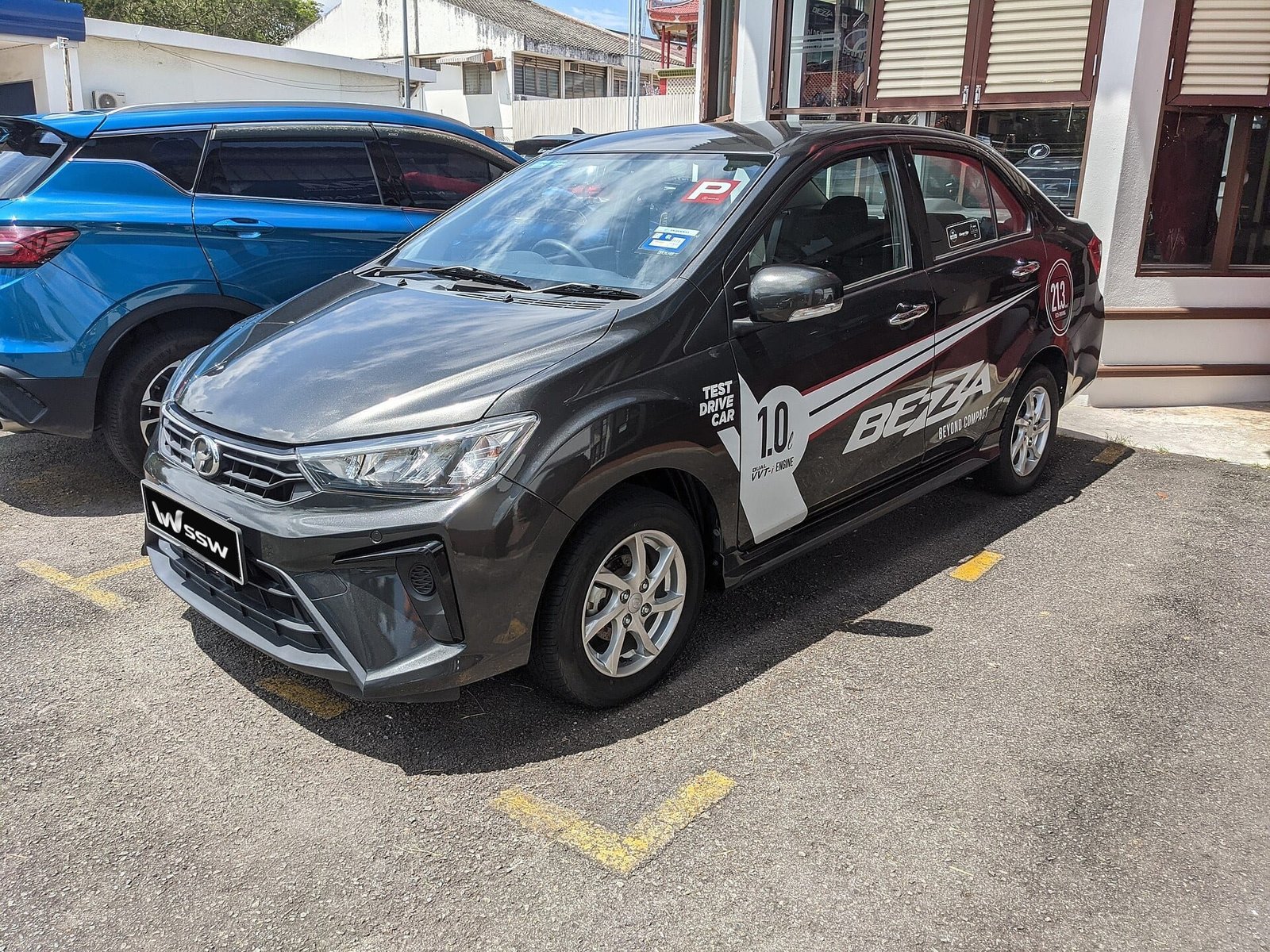 Granite grey Perodua Bezza 1.0 G sedan, front three-quarter view in a sunlit forecourt in Penang, Malaysia
