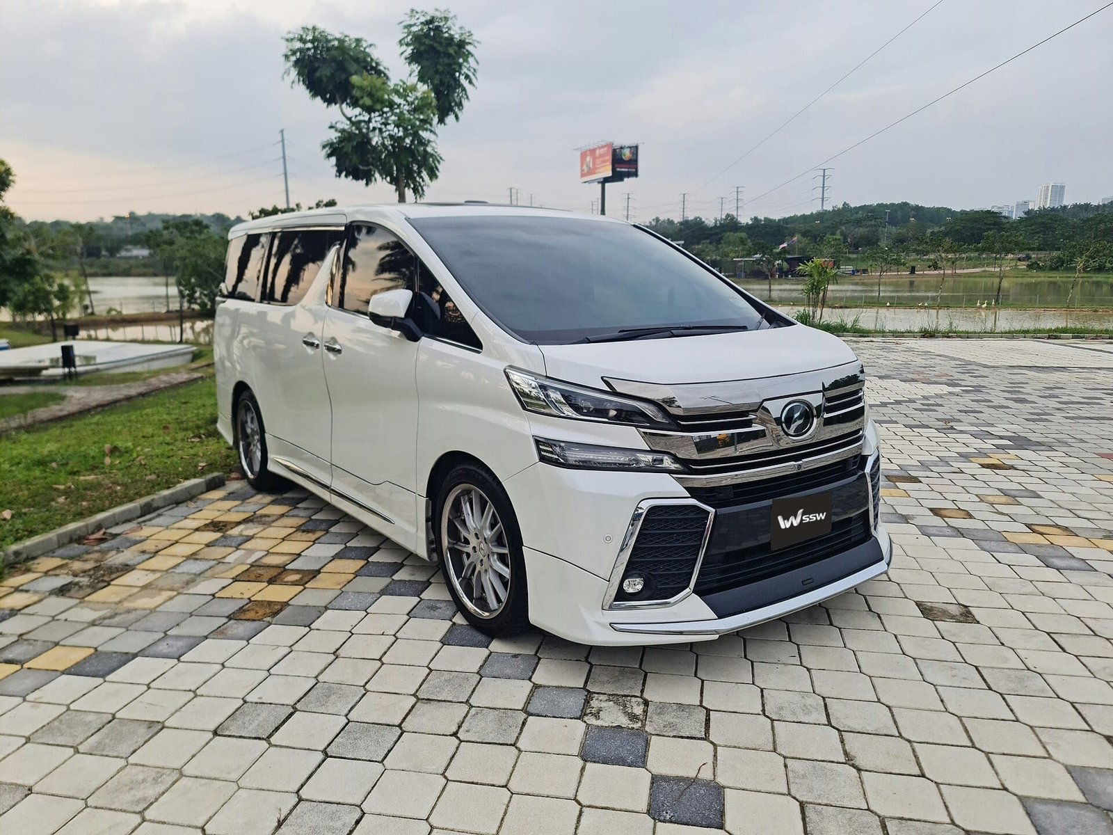 White Toyota Vellfire (AH30), front three-quarter view on a paved forecourt in Johor, Malaysia, with trees and a lake behind