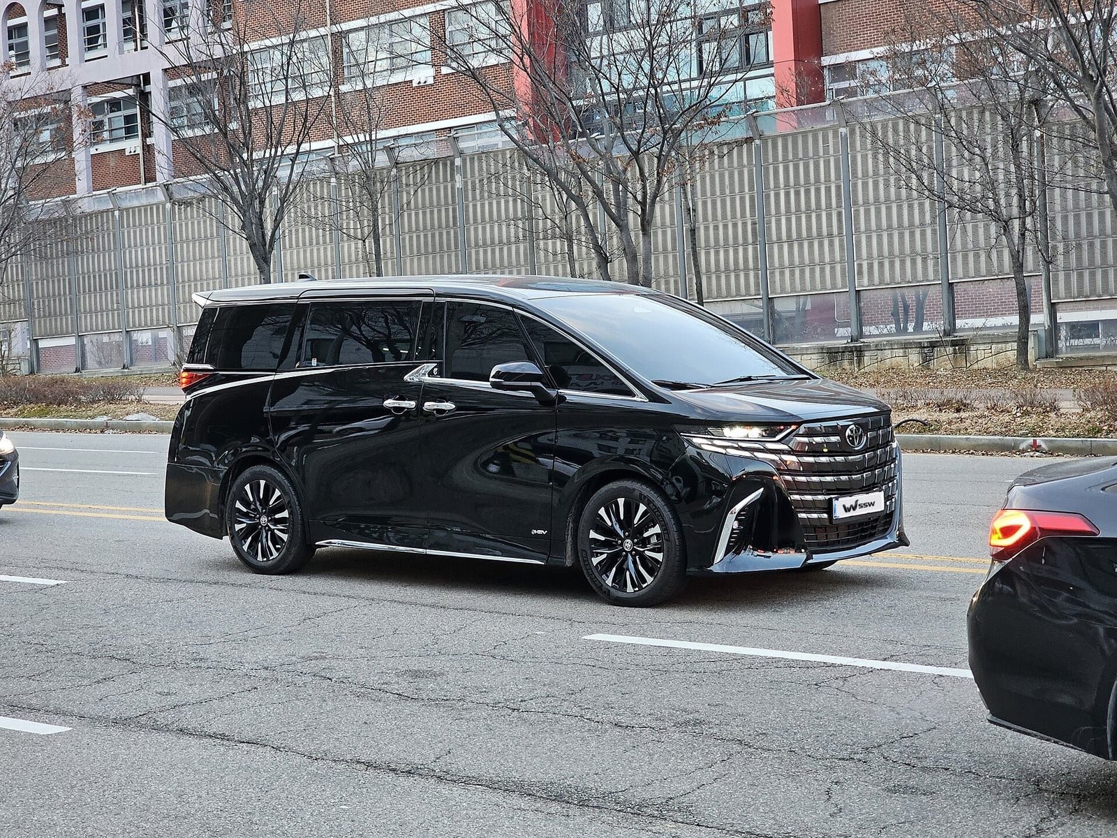Black Toyota Alphard (AH40) in side-front view on a road beside a fence and trees, in daylight
