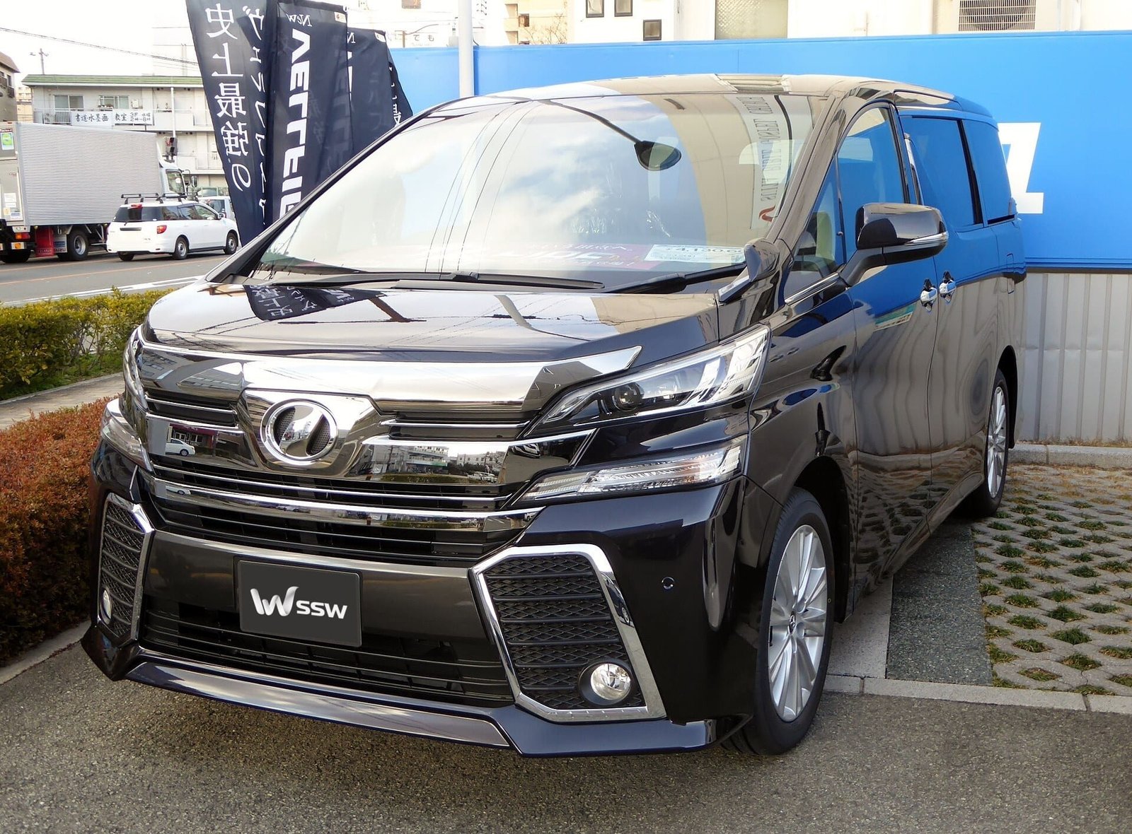 Black Toyota Vellfire ZA (AH30), front three-quarter view on a dealership forecourt in Japan, in daylight